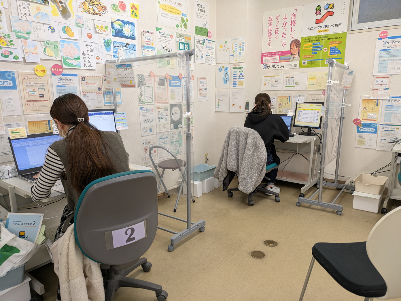 市民パソコン塾の教室内で、生徒がパソコン学習をしている様子。掲示物が多く、個別指導の雰囲気が伝わる教室内の写真。