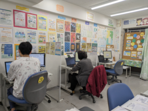 市民パソコン塾の教室内で、受講者がパソコンを操作している様子。壁に掲示物が並ぶ実際の教室風景。